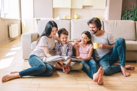 People are sitting on the floor. Boy has book. He reads it with sister and mom. Guy has a cup and look at book. He smiles.の写真素材