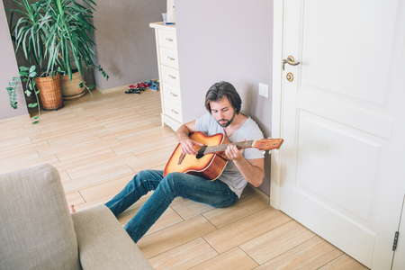 A picture of gy sitting alone on the floor and playing the guitar. He is leaning to the wall. Man is entertaining himself.の写真素材