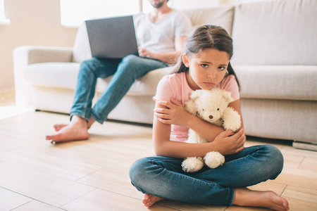 Small girl is sitting on floor. She is hugging her white bear toy. Girl looks to the right. She feels lonely. Her dad sits on sofa and work witth laptop.の写真素材
