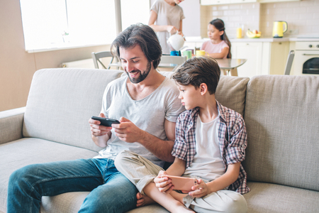 Positive man sits on sofa and plays on the phone. Boy has that phone as well but he looks at that his dad has. Girls are standing at table in kitchen and cooking.の写真素材