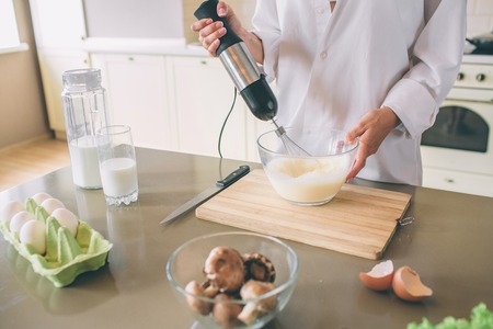 Cut view of woman mixing together eggs and milk with mixer. She holds bowl with one hand. Girl is...