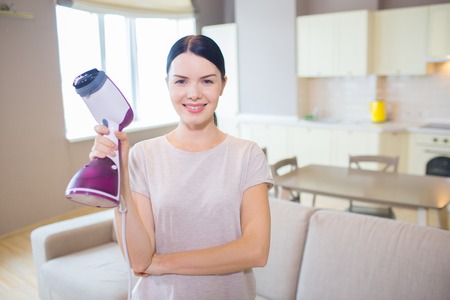 Pretty woman stands with small vacuum cleaner in hand and poses on camera.の写真素材