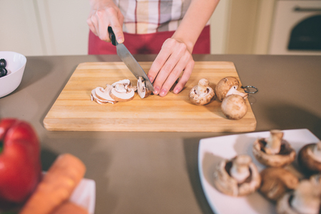 Close up of females hands cutting mushroom into pieces. She is doing that careful. Girl uses knife. There are plate with solid mushrooms and other vegetables.の写真素材