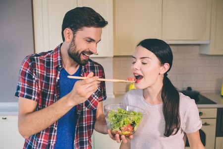 Horizontal portrait of couple standing at kitchen. Guy holds wooden spoon and feed girl. She keeps her mouth opened. They are tasing food they have cooked.の写真素材