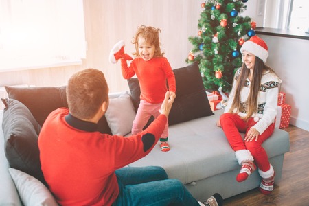 Merry Christmas and Happy New Year. Playful picture of happy child standing on couch and holding red hat. She looks at father and scream. Young dad hold his daughters hand. Woman look and smiles.の写真素材