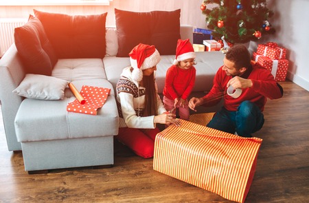 Merry Christmas and Happy New Year. Family sit on floor near big box of present. Young man shows his daughter how to tape edge of box. Girl looks at this. She is concentrated. Woman looks at daughter.の写真素材