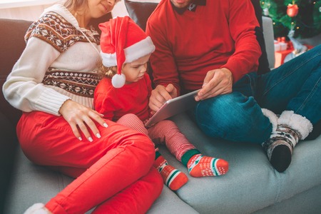 Merry Christmas and Happy New Year. Family Cut view of small girl sits between her parents. She holds tablet and points on it. Girl touches screen with finger. Man help to hold tablet.の写真素材