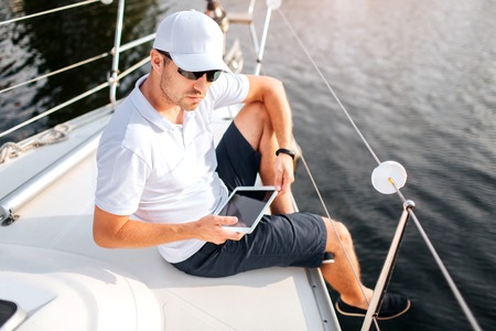 Picture of young man sits at edge of yacht board and hold tablet. It has dark screen. Man wears...