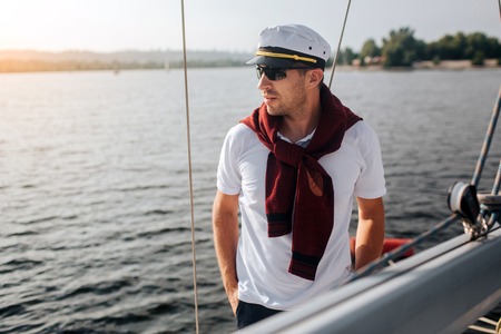 Sailor stand on his yacht. He keeps hands in pockets and look to left. Man is well-built and stylish. He is ready for trip.の写真素材