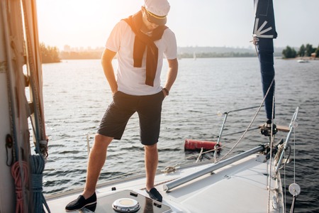 Another picture of oyung man stands on yacht. Hr keeps hands in pockets and look down. Young man has sweater on shoulders and cap on head. Also there are white shirt and dak short on his body. He poses.の写真素材