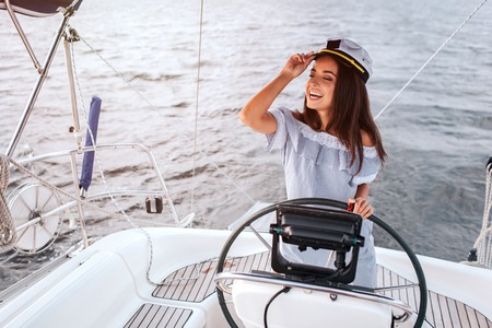 Beautiful model stands in front of rudder and uses it. She holds hand on cap and look to left. Woman is calm and peaceful. She is happy.の写真素材