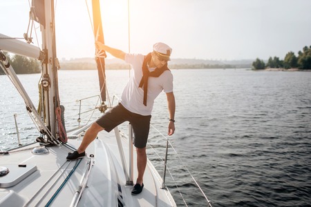 Beautiful icture of handsome and serious sailor standing on his yacht. He holds on tube and look at edge of boat. Young man poses. Sun goes down.の写真素材