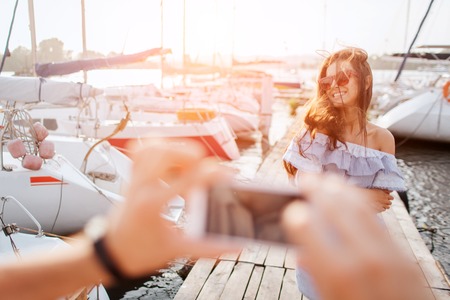 Beautiful and happy model stands and poses on camera. She smiles. Wind is blowing up her hair. Young woman wears sunglasses. Man takes a picture of brunette. They stand on pier. Sun goes down.の写真素材