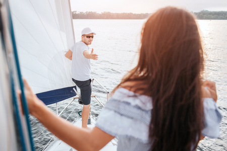 Young man in cap and glasses stands on bow of yacht and reaches to brunette with hand. He looks at her. She stands and looks at him. Model holds on tube.の写真素材