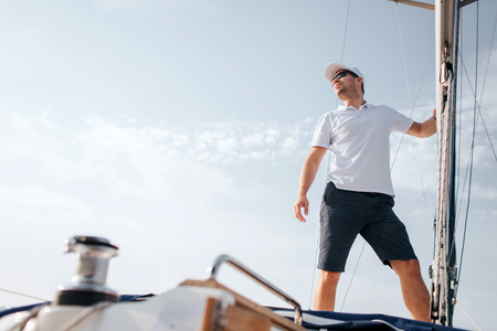 Young man stands on yacht board and looks forward. He holds on mast with hand. Young man poses. He wears white shirt and black shorts.の写真素材