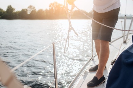 Cut view of man standing on yacht. He is at edge of it. Man holds white rope. He is workingの写真素材