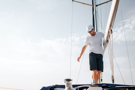 Handsome and confident man stands on yacht board and holds on big mast. He looks down and reaches with hand. Young man is serious and concentrated. He poses.の写真素材