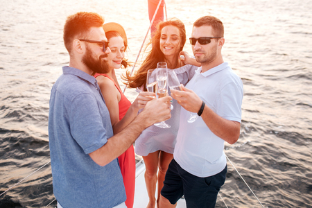 Four people standing close to each other. They touches with glasses of champaigne. Guys wear sunglasses. Young women smile and enjoy time.の写真素材
