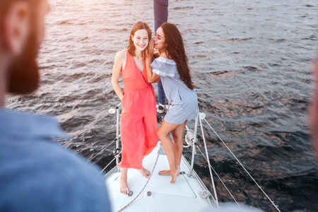 Beautiful young women stand together and pose. They wispering and laughing. Men stands adn look at them. Women are on bow of yacht.の写真素材