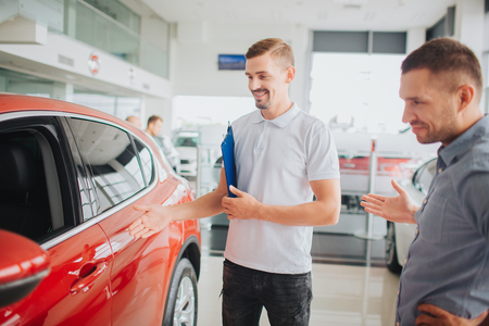 Car seller and customer stand by beautiful red car and look at it. They smile. Consultant holds plastic blue tablet. Customer point with his hand.の写真素材
