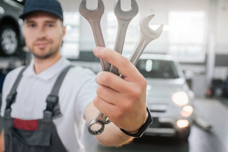 Young handsome man shows set of wrenches. He looks at them and smile. Guy wears uniform. He stands in front of white car. Young man smiles a bit.の写真素材