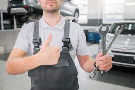 Picture of strong young man stands and shows big thumbs up. Also he holds two wrenches in left hand. There are tow cars behind him. One of them is on platform.の写真素材