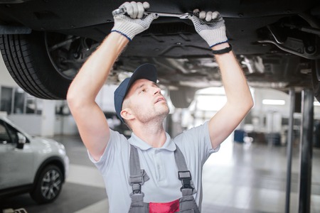 Young man in process of working underneath car. He looks up to right and hold big wrench with both hands. He is concentrated. Man works in garage.の写真素材