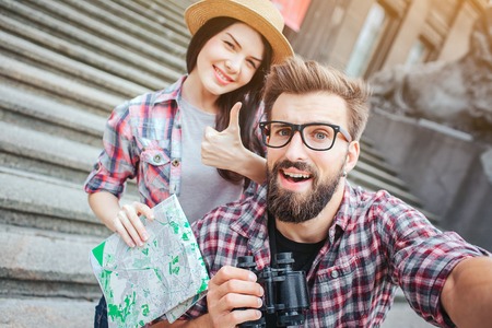 Happy and excited tourists stand on stairs and pose on camera. Bearded young man holds it. She shows big thumb up and keeps map in hand.の写真素材