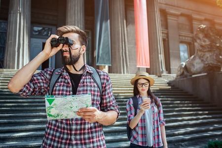 Serious and thoughtful man stand outside and look in binoculars. Also he holds map in hand. Female traveller stand behind him and look at phone she has in hands.の写真素材