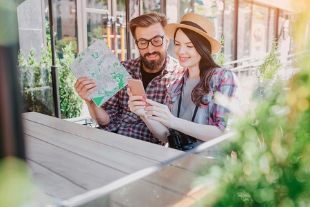 Lovely couple of trvellers sitting and looking at phone woman holds in hands. They smile. Young man holds map in hand. Tourists are surrounded by green plants.の写真素材