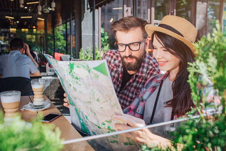 Interester young man and woman sit and look at map she holds. Woman smiles. Guy is serious. They have rest. There are coffee at table.の写真素材