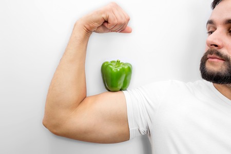 Nice picture of strong and well-built man holding green pepper on his muscular. He looks at it. Guy is calm and peaceful. Isolated on white background.の写真素材