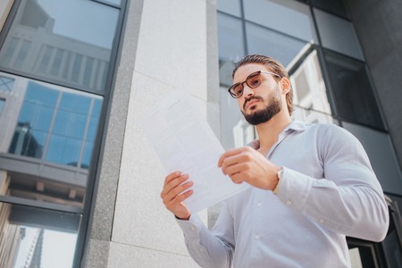 Picture of young stylish man stand and poses. He looks at white piece of paper through sunglasses. Guy is calm, peaceful and concentrated. He stands in front af glass building.の写真素材
