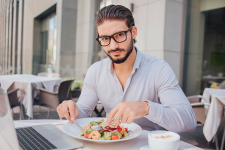 Young handsome man sits at table and has lunch. He looks at camera. Guy poses. He is ready to eat salad. There are laptop and cup of coffee at table.の写真素材