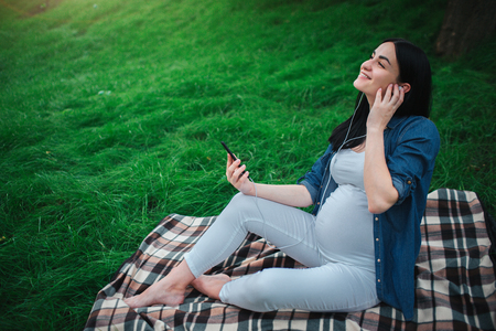 Portrait of a happy black hair and proud pregnant woman in a city in the background. She is sitting on a city bench. Expectant mother is listening to music in the park with an unborn childの写真素材