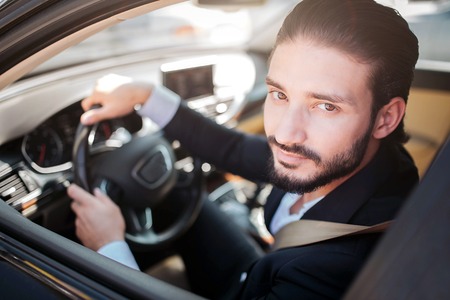 Satisfied and happy man sitting in car and posing. He looks at camera and smiles a bit. Bearded guy holds hands on steering wheel.の写真素材