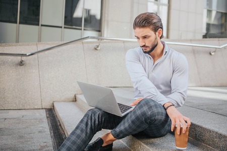 Young and stylish male person sits on stairs. He looks at laptop screen and reaches cup of coffee. Guy is busy. He works.の写真素材