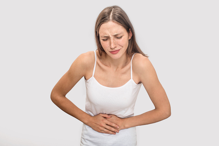 Blonde young woman with freckles stands and holds hands on stomach. She keeps eyes closed. Model suffering. Isolated on grey background.の写真素材