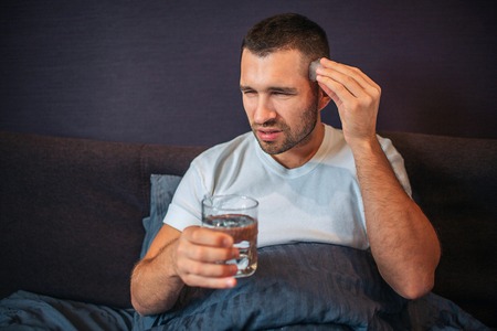 Young man sits on bed and shrinks. He has headache. Guy keeps one hand close to head and hold glass of water with another one. He is covered with blanket.の写真素材