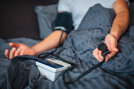 Cut view of man lying on bad with covered low part of body. He uses device for measuring blood...