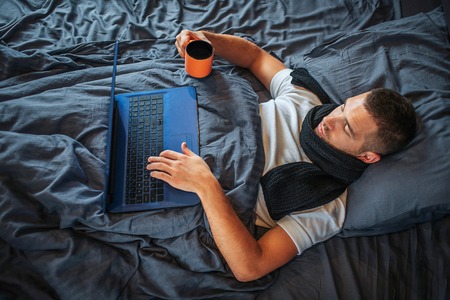 Sick young man works at home. He looks at screen of laptop and hold hand on keyboard. Guy holds cup of hot tea with another hand. He is calm and concentrated.の写真素材