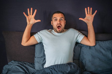 Wondered young man sits on bed and looks on straight on camera. He holds his hands up. Guy is scared and frightened.の写真素材