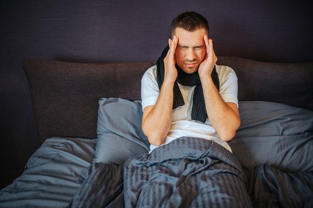 Young man sits on bed and holds hands close to forehead. He is suffering from headache. Pain is strong and terrible. Guy shrinks. He has scarf around neck.の写真素材