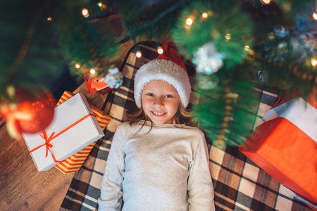 Beatufiul and positive small girl is lying under Christmas tree and looks on cmaera. She smiles. Girl wears Christmas hat. There are boxes with presents on both sides from her. She looks happy.の写真素材