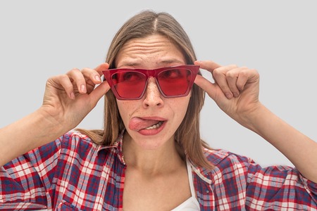 Funny young woman stands close and looks to left through red sunglasses. She holds it with both hands and shows tongue. Woman is playing role. Isolated on grey background.の写真素材