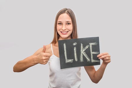 Very positive and attractive young woman looks on camera and smiles. She holds plate with written word like on it. Also model shows her big thumb up. Woman is happy. Isolated on grey background.の写真素材