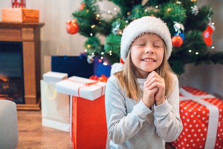 Blonde girl stands and prays. She keeps hands together and keeps eyes closed. Girl wishing. There are boxes of presents besides Christmas tree behind child.の写真素材