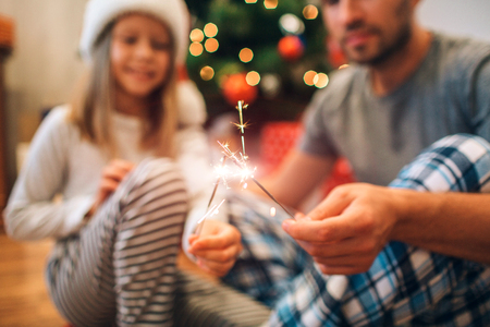 Cut view of small family holding together Bengal lights sticks with sparkling lights. They look at it. People sits on floor in decorated room.の写真素材