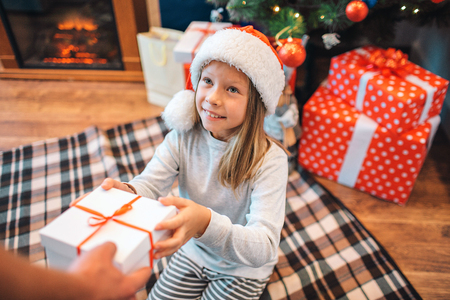 Cheerful girl takes box with gift and looks at adult that gives it to her. She smiles a bit. Girl is thankful. She sits on blanket. There are another surprises and Christmas tree behind her.の写真素材