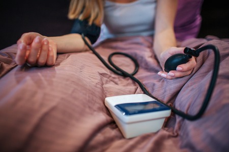 Cut view of young woman sitting on bed and mesuring blod pressure with special tool. She squizes...